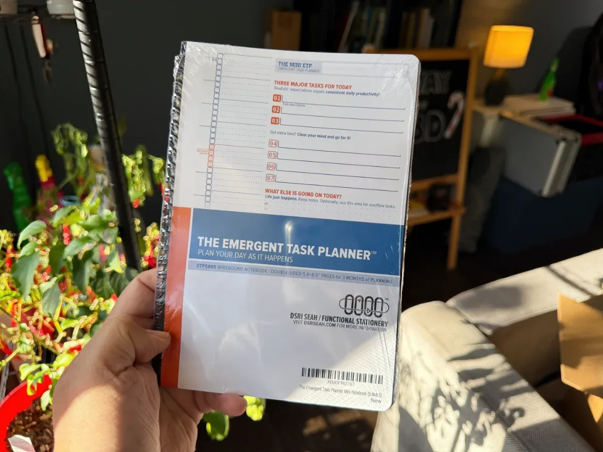 A shrinkwrapped Mini ETP Notebook held in the hand to show its size, against a backdrop of Sri's messy living room (full size image)