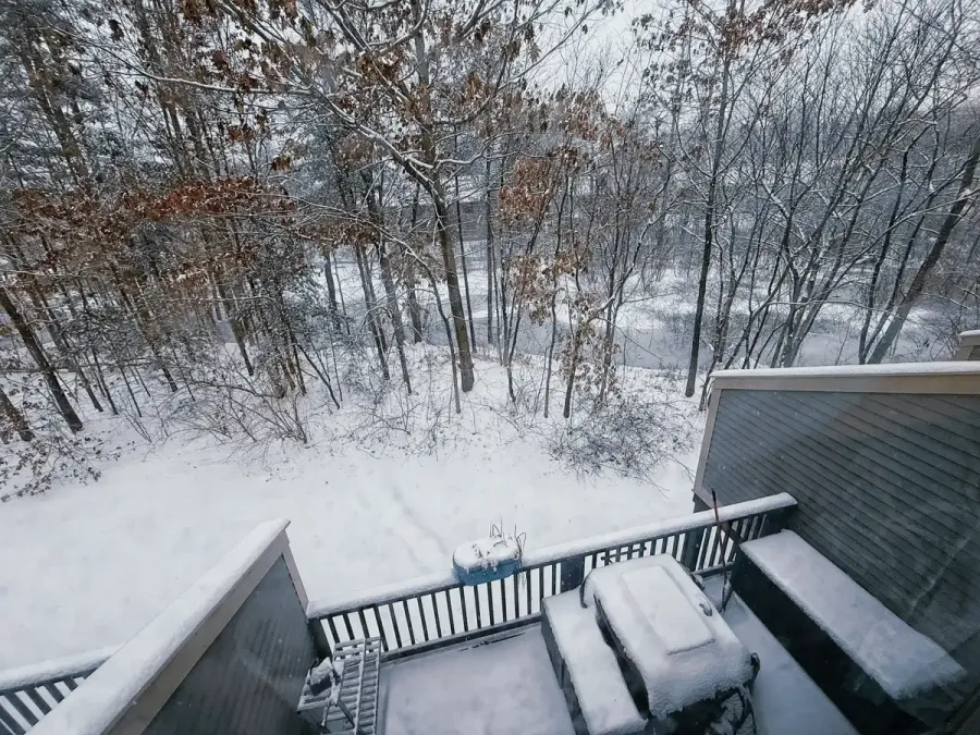 The view of a snowy back deck taken from the second story of a condominium, looking over an icy creek with sparse pine tree cover