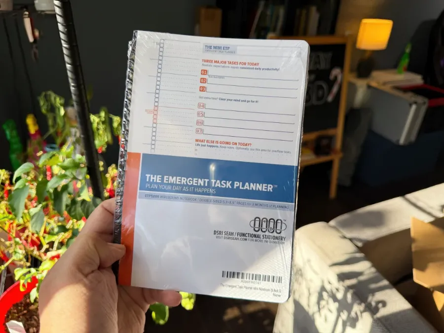 A shrinkwrapped Mini ETP Notebook held in the hand to show its size, against a backdrop of Sri's messy living room